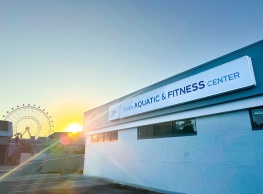 A building with a sign that reads “Rosen Aquatic and Fitness Center” at sunset, with a sun glare and a Ferris wheel in the background.