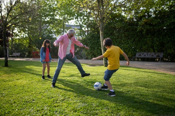 Alt text: Outside, a boy kicks a soccer ball toward an older man and a young girl standing behind him. 