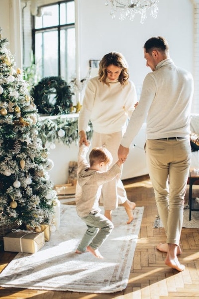 A man and woman in neutral colors dance in a circle holding hands with a young boy in a neutrally decorated room with a Christmas tree and other holiday decor. 