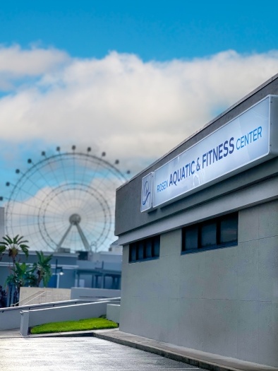 The side of a building with a sign that reads “Rosen Aquatic and Fitness Center” with a blue sky and a large Ferris wheel in the background.