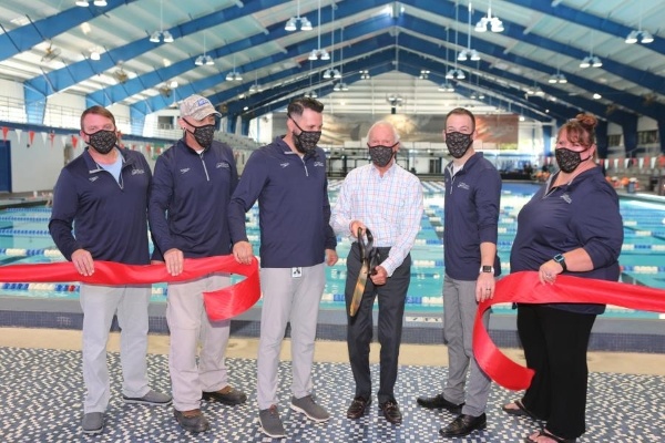 Six people in masks, five in matching navy polos and one older gentleman in a light button down holding large scissors (Harris Rosen) stand in front of a cut red ribbon. 