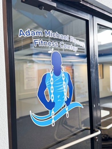 A decal on a window that reads "Adam Michael Rosen Fitness Center" with a blue silhouette of a muscular man holding a chain around his neck. 