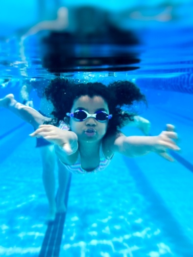 A little girl with goggles on swims underwater in a swimming pool. 