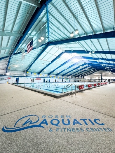 An indoor pool beneath a large ceiling with blue beams, and the Rosen Aquatic & Fitness Center logo painted on a concrete pool deck in the foreground.