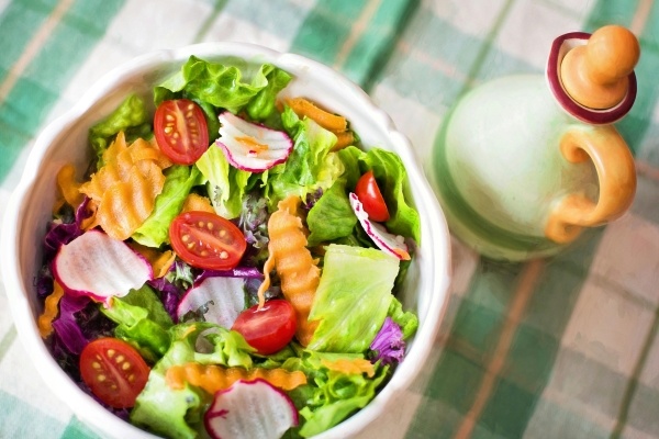 A bowl with chopped lettuce, tomatoes, radishes, and carrots against a green and yellow plaid tablecloth next to a ceramic bottle. Ordering a salad is a great healthy choice at a restaurant. 