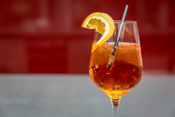 A red-orange cocktail in a wine glass with a black straw and an orange slice against a red and gray background. 