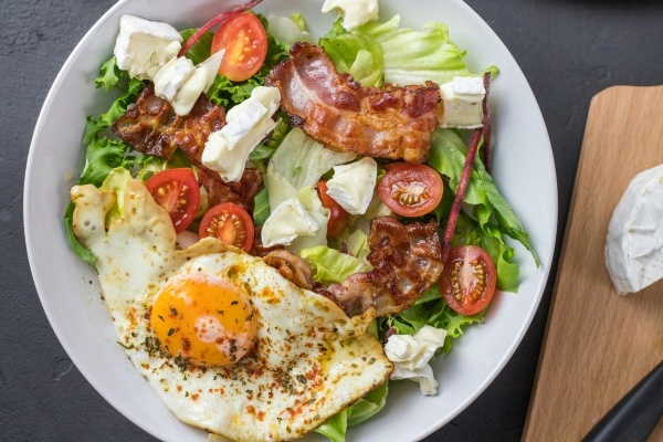 A salad in a white bowl topped with tomatoes, bacon, a fried egg, and thick pieces of brie cheese. 