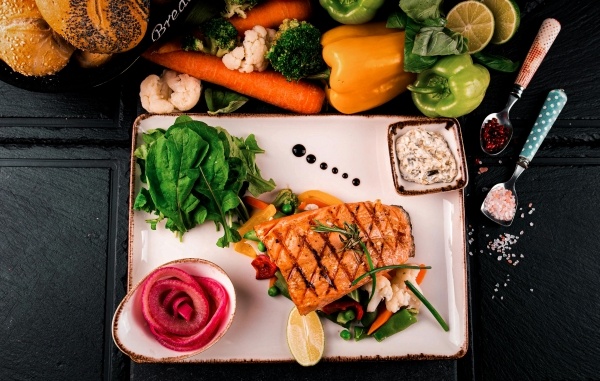 A white plate with a piece of seared salmon over a bed of veggies next to a flower-shaped pink garnish, spinach, and a dish with sauce against a black background and next to a display of fresh veggies. 