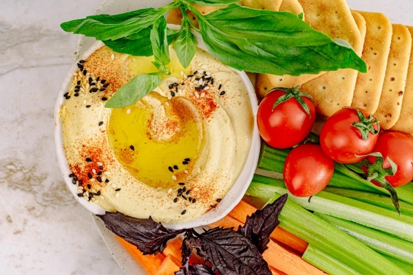 A bowl of hummus topped with olive oil and black and white sesame seeds next to a plate of tomatoes, pita, celery, carrots, and green and purple basil. 