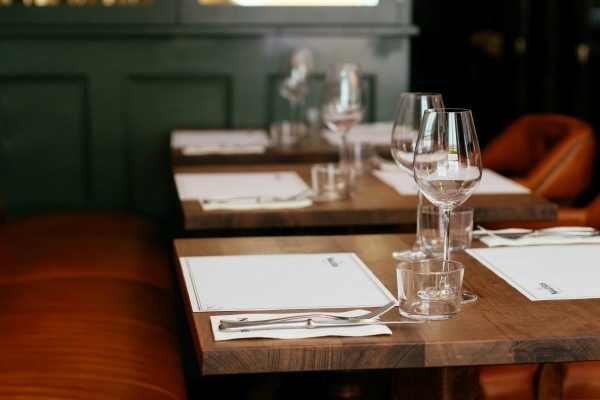 A row of three wooden tables with rust-colored chairs, set with wine glasses, menus, and silverware at a restaurant. 