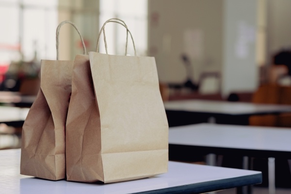 Two brown paper bags with handles sitting on a white table in a room of similar tables. 